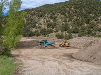 two yellow bulldozers work on sand and gravel in a mountain valley while another one looks at it