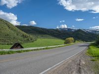 the empty road is going through a valley with mountains and grass in the background and two houses on both sides