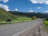 the empty road is going through a valley with mountains and grass in the background and two houses on both sides