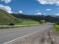 the empty road is going through a valley with mountains and grass in the background and two houses on both sides