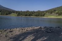 a rocky shore next to the water in a lake surrounded by mountains and trees on a clear day