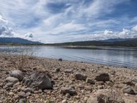 a lake with rocks on the shore and mountains in the background along side it and snow capped hills