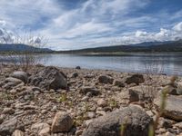 a lake with rocks on the shore and mountains in the background along side it and snow capped hills