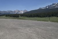 a dirt field and road near pine trees and mountain tops against blue sky with wispy clouds