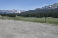 a dirt field and road near pine trees and mountain tops against blue sky with wispy clouds