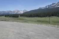 a dirt field and road near pine trees and mountain tops against blue sky with wispy clouds