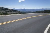 an empty asphalt highway with yellow lines down the middle of it next to mountains and blue skies