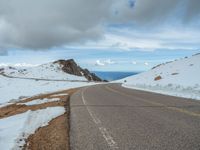 an empty road with snow and mountains in the back ground under cloudy skies on a sunny day