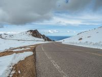 an empty road with snow and mountains in the back ground under cloudy skies on a sunny day