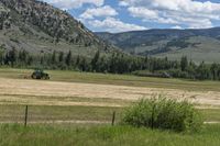 a green tractor is driving through a grassy field with mountains behind it and trees in the foreground