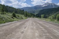 dirt road surrounded by mountain with evergreen trees in distance and blue sky with white clouds above