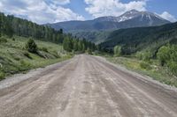 dirt road surrounded by mountain with evergreen trees in distance and blue sky with white clouds above