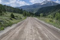 dirt road surrounded by mountain with evergreen trees in distance and blue sky with white clouds above