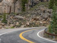 the curved road curves in front of a mountain area with pine trees behind it,