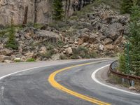 the curved road curves in front of a mountain area with pine trees behind it,