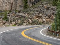 the curved road curves in front of a mountain area with pine trees behind it,