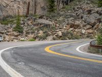 the curved road curves in front of a mountain area with pine trees behind it,