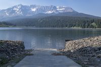 a skateboarder stands on a dock next to a mountain lake with snowy mountains in the background