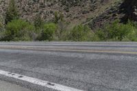 an empty street near some mountain side with yellow lines and some trees behind it to the left