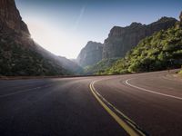 an empty mountain road going in between high mountains with a blue sky and sun reflecting in the background