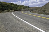 an empty, winding road runs in front of mountains and trees under a cloudy sky