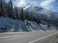 a long highway going through some evergreen trees and snowy hills and mountains behind it and snow on the ground