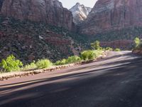 a truck drives on the road towards some mountain range and canyons behind it, with a stone - clad fenced pathway and trees on either side