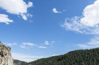 a kite flies through a blue sky near a cliff with trees in the background and a hill covered with rocks