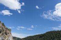 a kite flies through a blue sky near a cliff with trees in the background and a hill covered with rocks