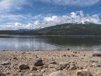 a lake with rocks on the shore and mountains in the background along side it and snow capped hills