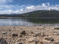 a lake with rocks on the shore and mountains in the background along side it and snow capped hills