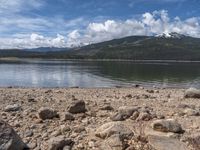 a lake with rocks on the shore and mountains in the background along side it and snow capped hills