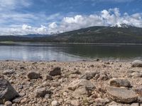 a lake with rocks on the shore and mountains in the background along side it and snow capped hills