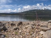 a lake with rocks on the shore and mountains in the background along side it and snow capped hills