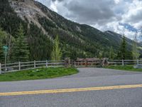 road in the mountains with grass and trees and a fence surrounding it on a bright day