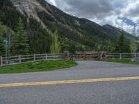 road in the mountains with grass and trees and a fence surrounding it on a bright day