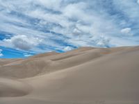 a lone horse standing on top of a sand covered hillside beneath a blue cloudy sky