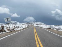 snow covers the roadway and snowy mountains on a sunny day, with a yellow warning sign in front of it