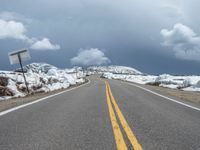 snow covers the roadway and snowy mountains on a sunny day, with a yellow warning sign in front of it