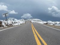 snow covers the roadway and snowy mountains on a sunny day, with a yellow warning sign in front of it