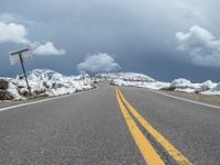 snow covers the roadway and snowy mountains on a sunny day, with a yellow warning sign in front of it