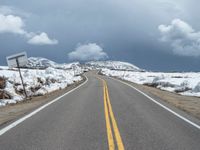 snow covers the roadway and snowy mountains on a sunny day, with a yellow warning sign in front of it