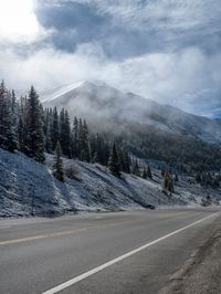 a car is parked along a mountain road with trees in the snow on one side and snowy mountains in the other