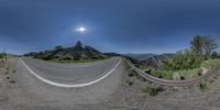 three fisheye image with a bend road between two mountains in the distance, with a man riding a skateboard below