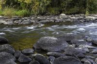 rocks and water on the shore of a river in an autumn forest setting, with foliage on either side