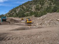 two yellow bulldozers work on sand and gravel in a mountain valley while another one looks at it