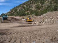 two yellow bulldozers work on sand and gravel in a mountain valley while another one looks at it