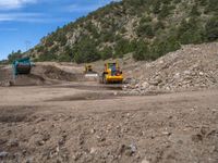 two yellow bulldozers work on sand and gravel in a mountain valley while another one looks at it