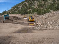 two yellow bulldozers work on sand and gravel in a mountain valley while another one looks at it