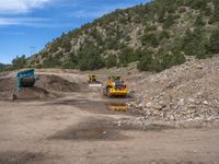 two yellow bulldozers work on sand and gravel in a mountain valley while another one looks at it